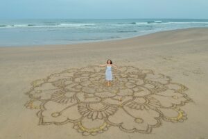 a large mandala painted on the sand on the ocean with a girl in the middle boho style.