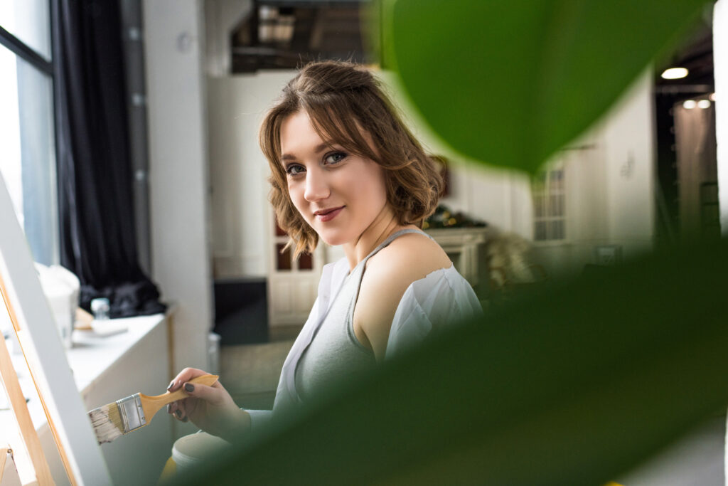 Young artistic girl looking at camera through leaves while painting in light studio